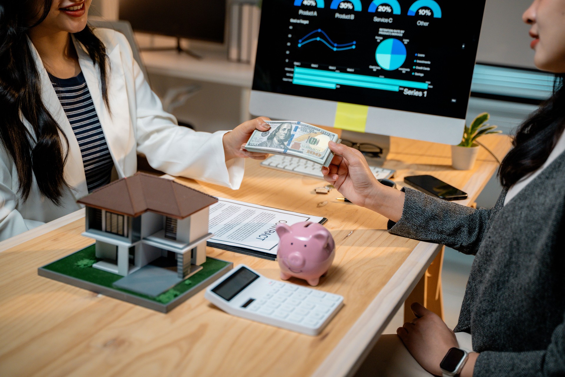 Real estate agent receiving cash down payment from client for house purchase, with house model, contract, piggy bank and calculator on desk